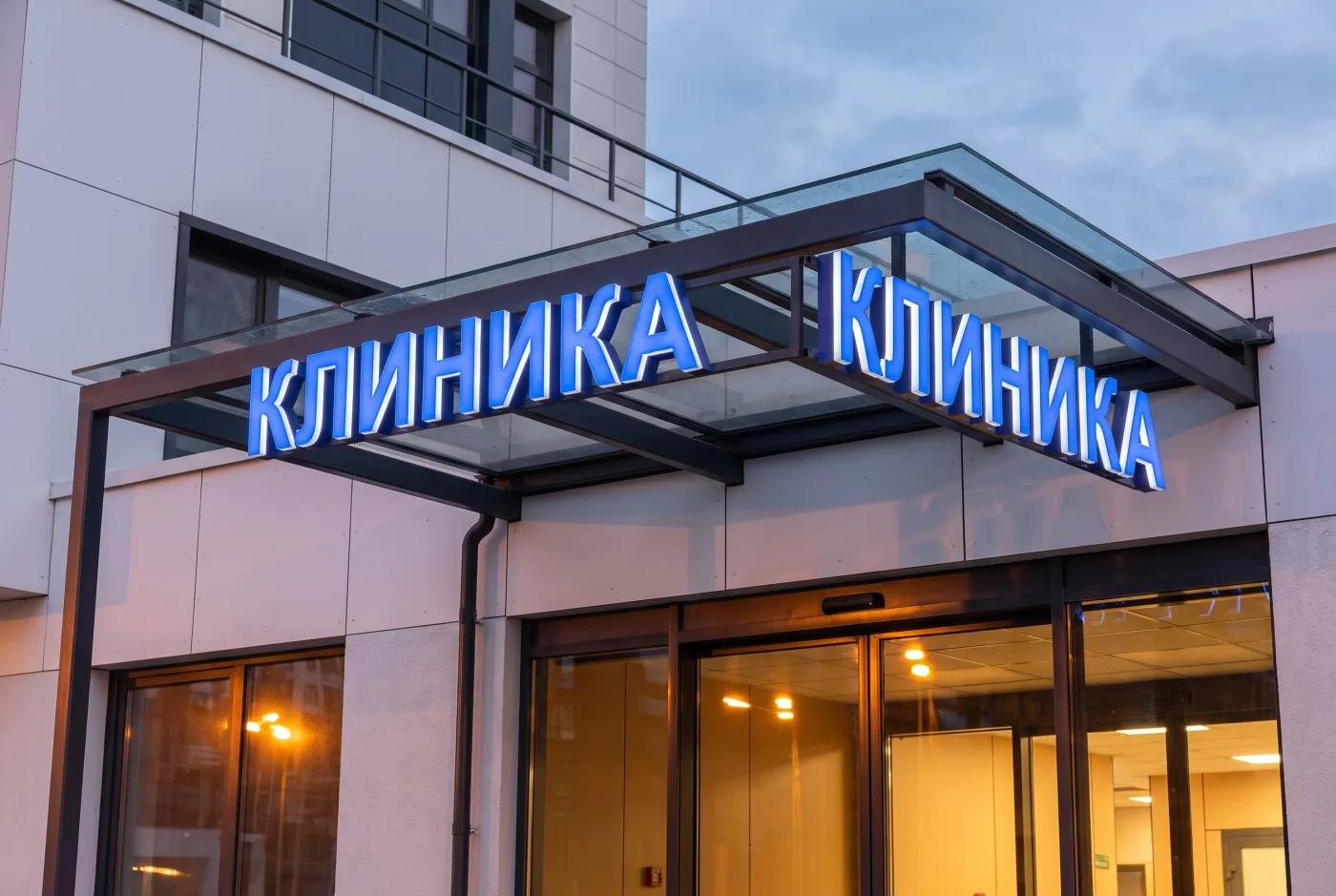 Подсвеченная вывеска КЛИНИКА на фасаде медицинского центра крупным планом — кириллические световые буквы, голубой LED-контур, навес над входной группой