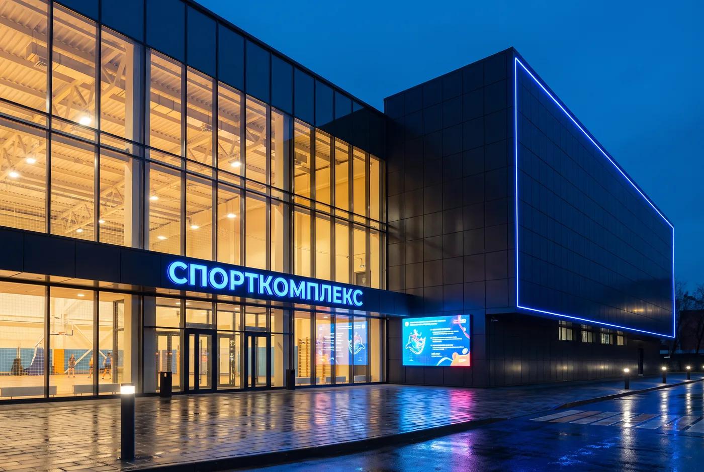 Вечерний облик с подсветкой Спортивный комплекс в вечерней подсветке — светящиеся изнутри витражи зала, контурная LED-линейка по объёму, медиатабло, площадь перед входом, AI-рендер